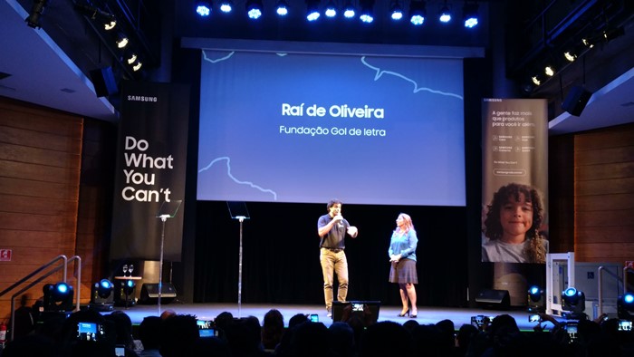 Imagem do palco, onde estão o ex-jogador Raí e Andréa Mello. Na tela azul em cima do palco está escrito "Raí de Oliveira, Fundação Gol de Letra"