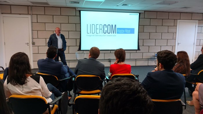Foto de Pedro Herz em pé, de frente para a plateia. Na tela, está projetado a marca do Lidercom, ao lado da marca, encontra-se uma caixa azul escrito "Happy hour".